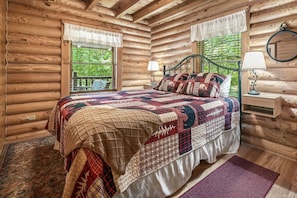 A log cabin bedroom with a bed, a nightstand, a lamp, and a window with a view of greenery.