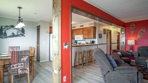 A dining area with a wooden table and chairs, a kitchen with wooden cabinets, and a living room with a gray sofa and a red accent wall. - Summit 304B  3 Bedrooms 2 Full Baths (Snowshoe)