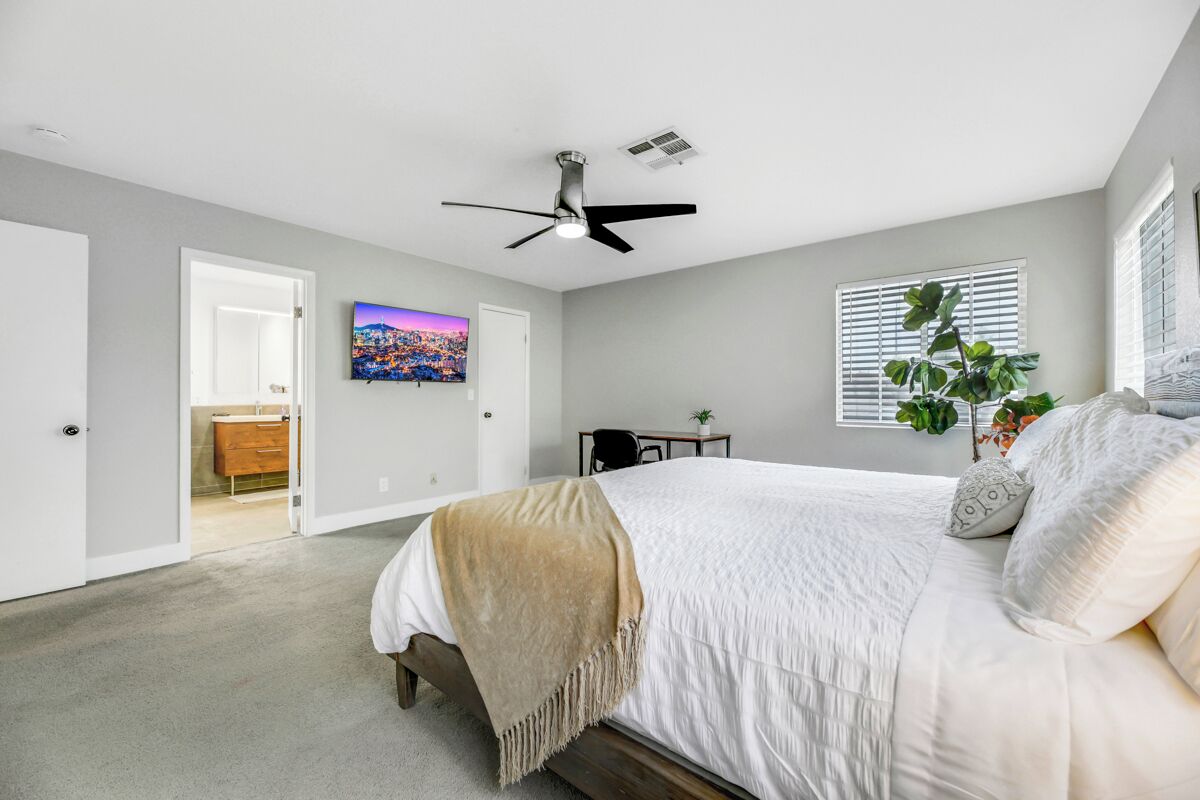 A bedroom with a bed, a ceiling fan, a window with blinds, a plant, and a painting on the wall.