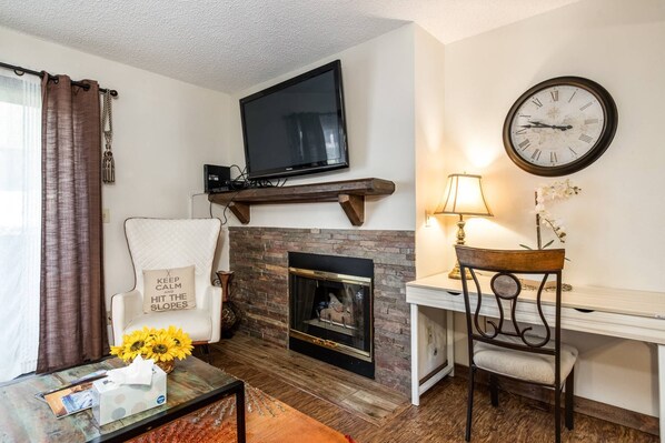Una sala de estar con chimenea, un televisor montado encima, un escritorio con una silla y un reloj en la pared.