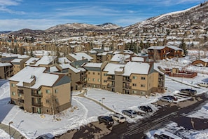 A residential area with multi-story apartment buildings, a parking lot, and a mountainous backdrop. - August Monthly Rates! Summer Pool + Hot Tub-Grill-W/D in Unit-Walk to Mtn/Steps to Free Town Bus (Steamboat Springs)