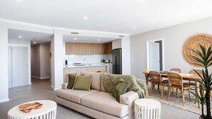 Un salon moderne avec un canapé beige, une table à manger entourée de chaises en osier, et une cuisine en arrière-plan.