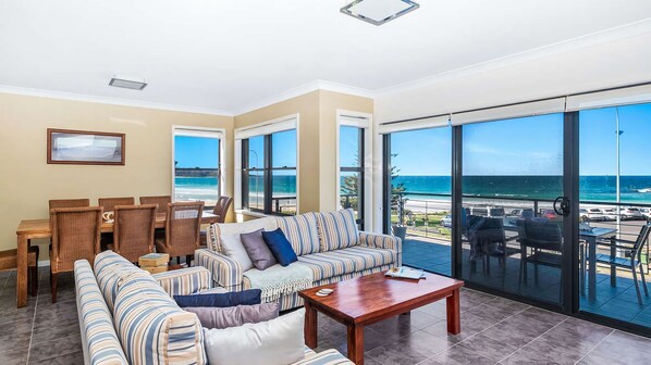 A spacious living room with a large sofa, a wooden coffee table, and a dining area with a wooden table and chairs. The room has large windows offering a view of the beach and ocean.