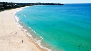 A wide sandy beach with clear turquoise water, a few people walking along the shore, and a distant shoreline with buildings.