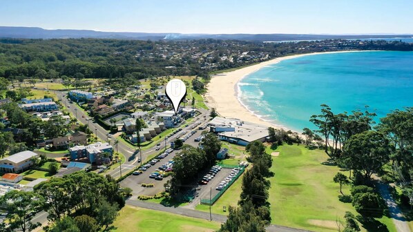 An aerial view of a coastal area with a beach, a parking lot, and residential buildings. - Beachfront Four - Remarkable apartment with breathtaking views (Mollymook)
