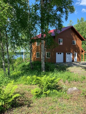 A two-story wooden house with a red roof, surrounded by lush greenery and a lake in the background.