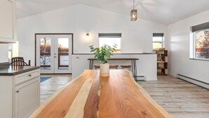 A modern kitchen with a large wooden table, white cabinetry, and a view of the outdoors through a door. - DENALI PENTHOUSE, lakefront near Talkeetna and Denali State Park  (Trapper Creek)