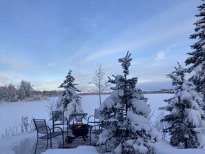 A snow-covered outdoor seating area with a fire pit, surrounded by snow-laden trees and a clear sky.