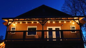 A two-story wooden cabin with a balcony, string lights, and a dark sky background. - DENALI PENTHOUSE, lakefront near Talkeetna and Denali State Park  (Trapper Creek)