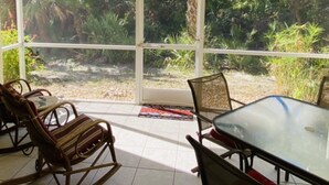 Een overdekt terras met een glazen tafel en stoelen, omringd door weelderige begroeiing en palmbomen.