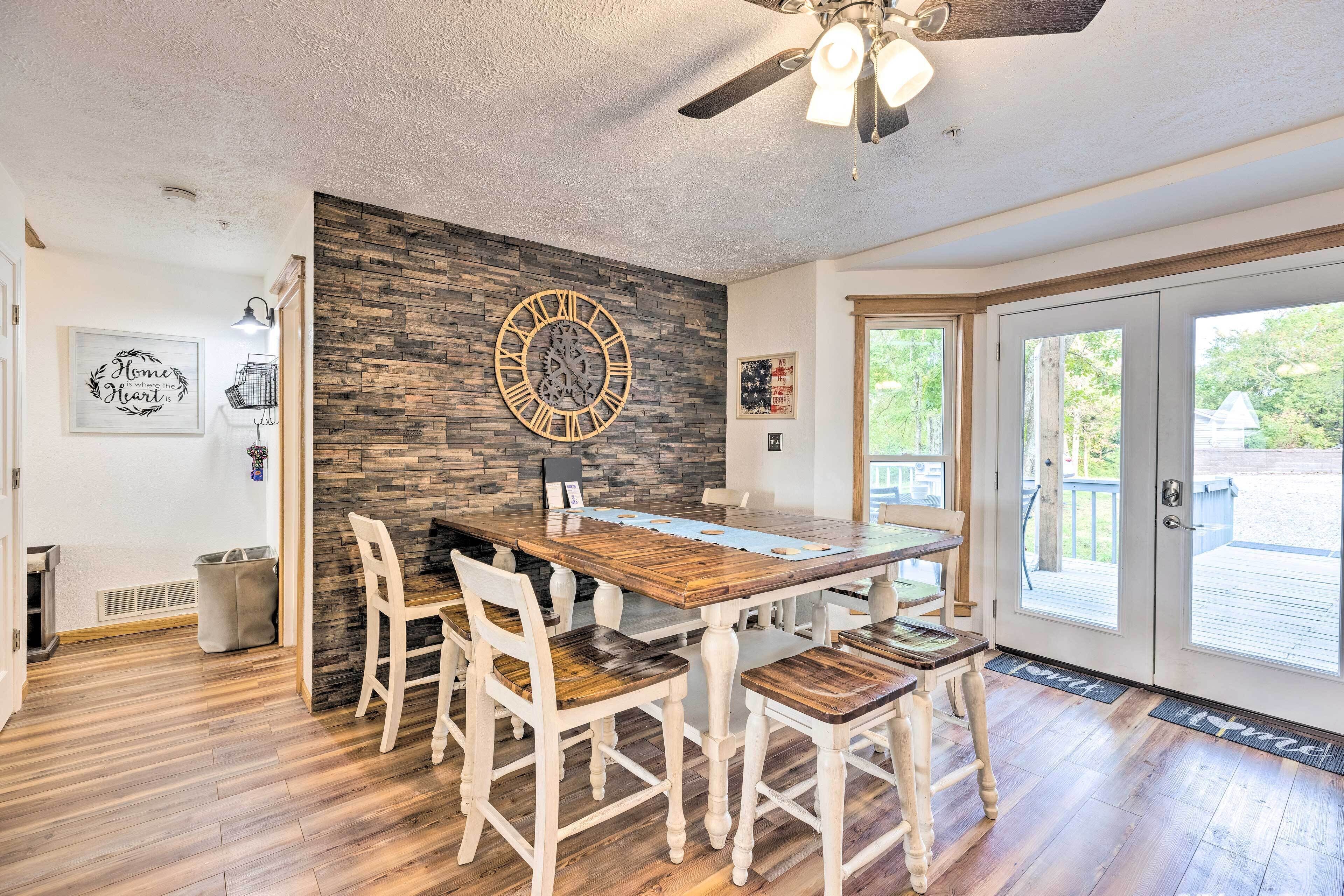 Un comedor con una mesa de madera y sillas blancas, una pared de piedra y un reloj grande.