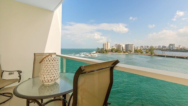 Un balcón con mesa y sillas de vidrio, con vistas al mar azul y al horizonte de la ciudad.