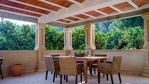 A covered patio with a table and chairs, surrounded by greenery and orange trees.
