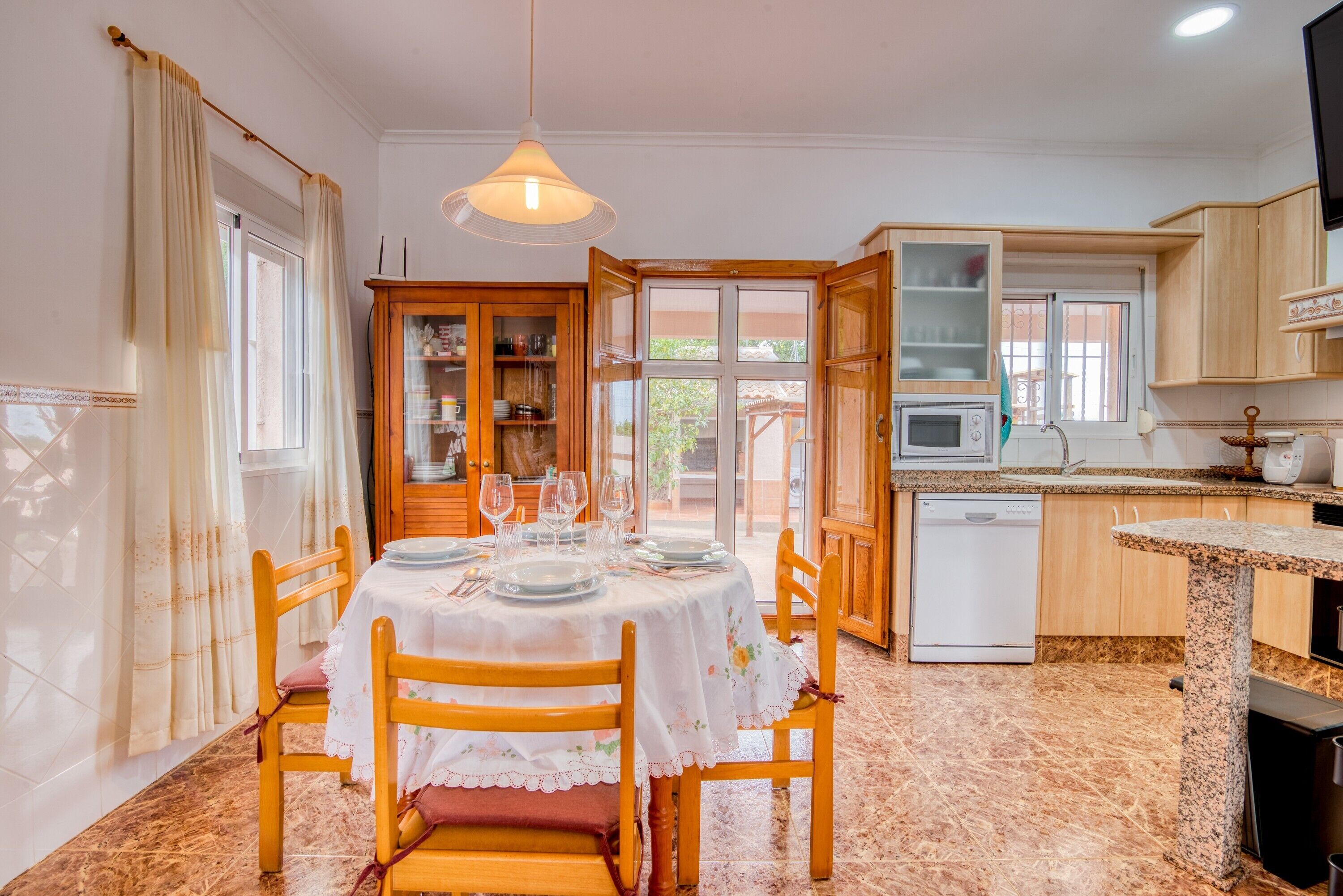 Un comedor con una mesa dispuesta para cuatro, sillas de madera, un armario con puertas de vidrio, un microondas y un lavavajillas.