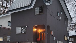 Ein modernes, zweistöckiges Haus mit dunkler Fassade, einem geneigten Dach und einem sichtbaren Eingang mit beleuchtetem Innenraum.