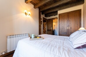 A bedroom with a bed, a radiator, wooden ceiling beams, a wall-mounted light fixture, and a door leading to another room.
