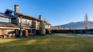 Une maison en pierre à deux étages, dotée d’un balcon, d’une pelouse et avec des montagnes en arrière-plan.