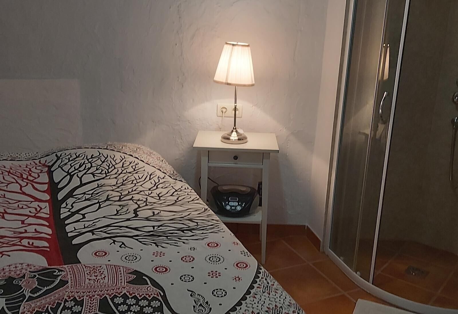 Un dormitorio con cama, una mesita de noche con lámpara, un estante pequeño y una puerta corrediza de vidrio que da a un baño.