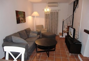 Un salon comprenant un canapé, un pouf, un fauteuil blanc, une lampe, un lustre et un téléviseur posé sur un meuble.