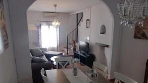 A dining area with a table, chairs, a chandelier, a staircase, and a television in the background.