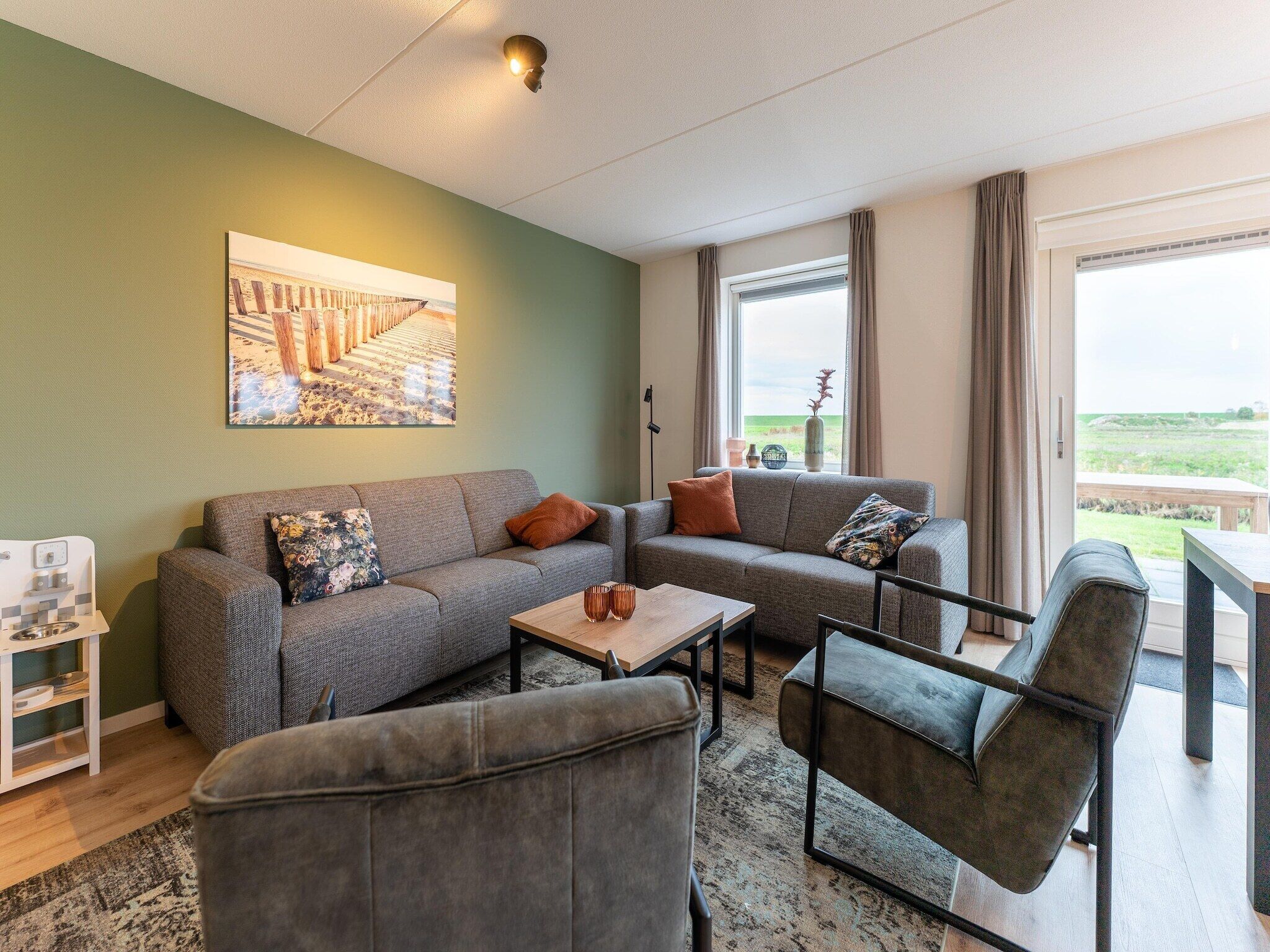 A modern living room with a grey sofa set, a coffee table, and a grey armchair. There is a painting on the wall, a window with curtains, and a view of the outdoors.