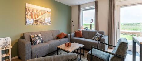 A modern living room with a grey sofa set, a coffee table, and a grey armchair. There is a painting on the wall, a window with curtains, and a view of the outdoors.