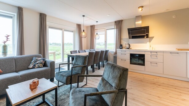 En moderne stue med sofa, lænestole, sofabord og spiseområde med bord og stole. Der er et køkken med mikrobølgeovn og skabe.