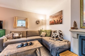 Een gezellige woonkamer met een hoekbank, een houten salontafel, een open haard en een groot raam met gordijnen.