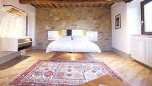 Ein Schlafzimmer mit Steinwand, Holzbalken, einem Bett mit floralem Überwurf, Nachttischlampen, einem hängenden Kleiderständer und einem gemusterten Teppich.