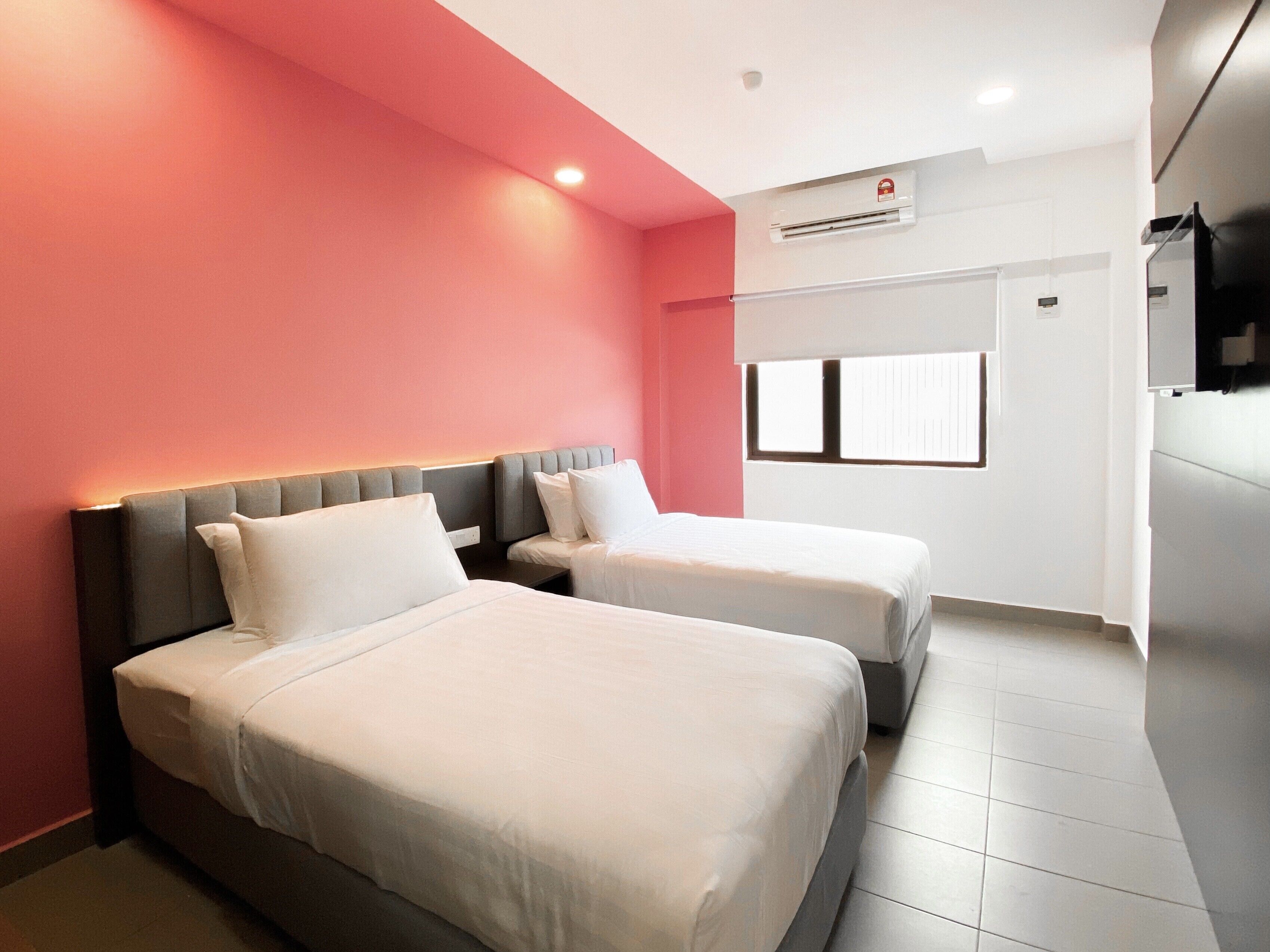 A hotel room with two beds, a red accent wall, and a window with blinds.