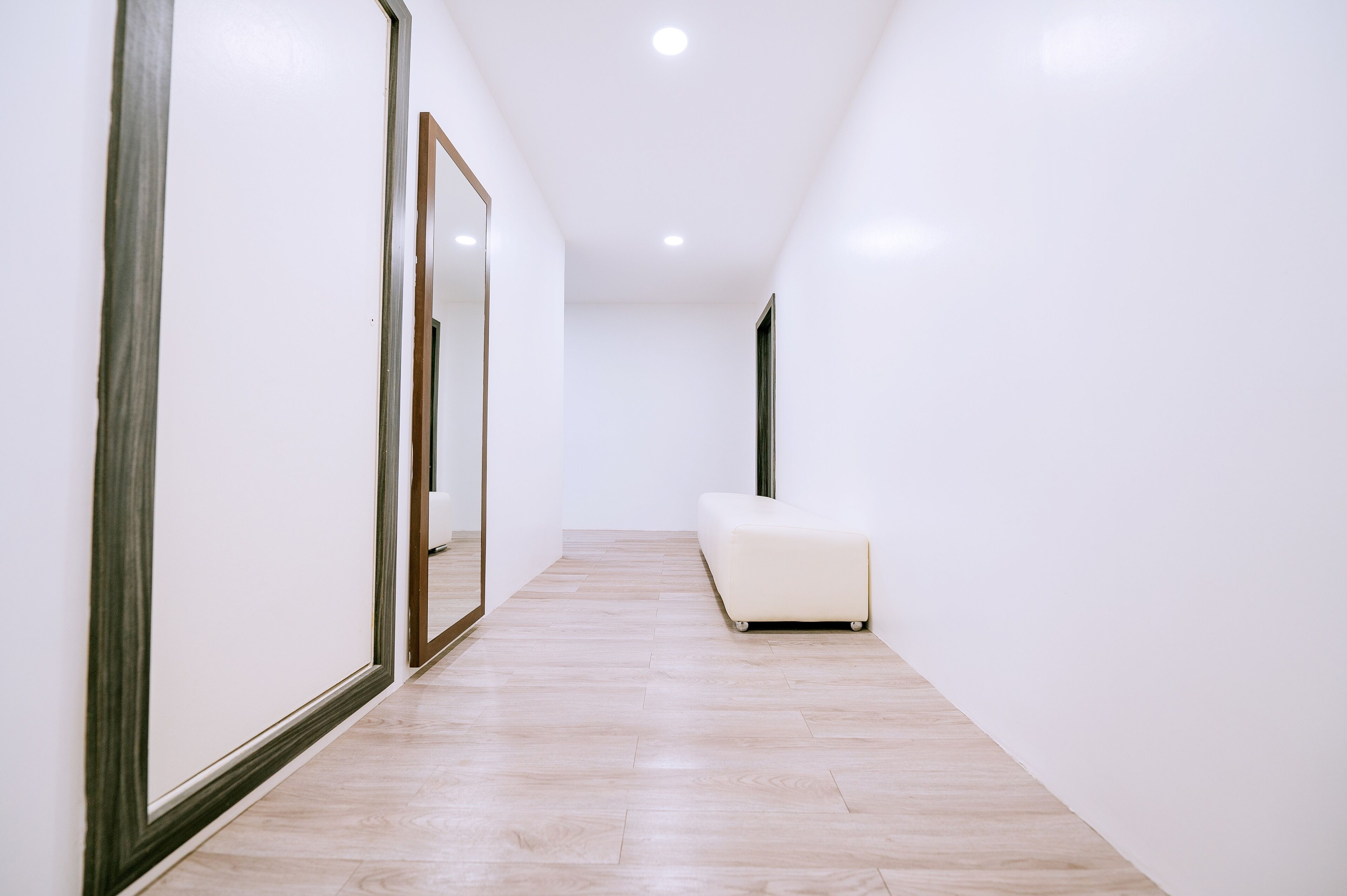 Un pasillo estrecho con un banco blanco, suelo de madera y un espejo grande en la pared izquierda.