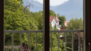 A view through a window with a white railing, featuring a house with a red roof surrounded by lush greenery and mountains in the background. - Apartment in the heart of Bled with views (Bled)
