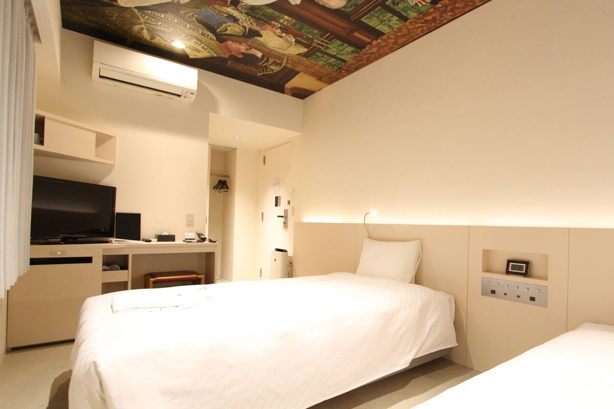 Habitación de hotel con dos camas, un escritorio y un televisor, y un techo con diseño decorativo.