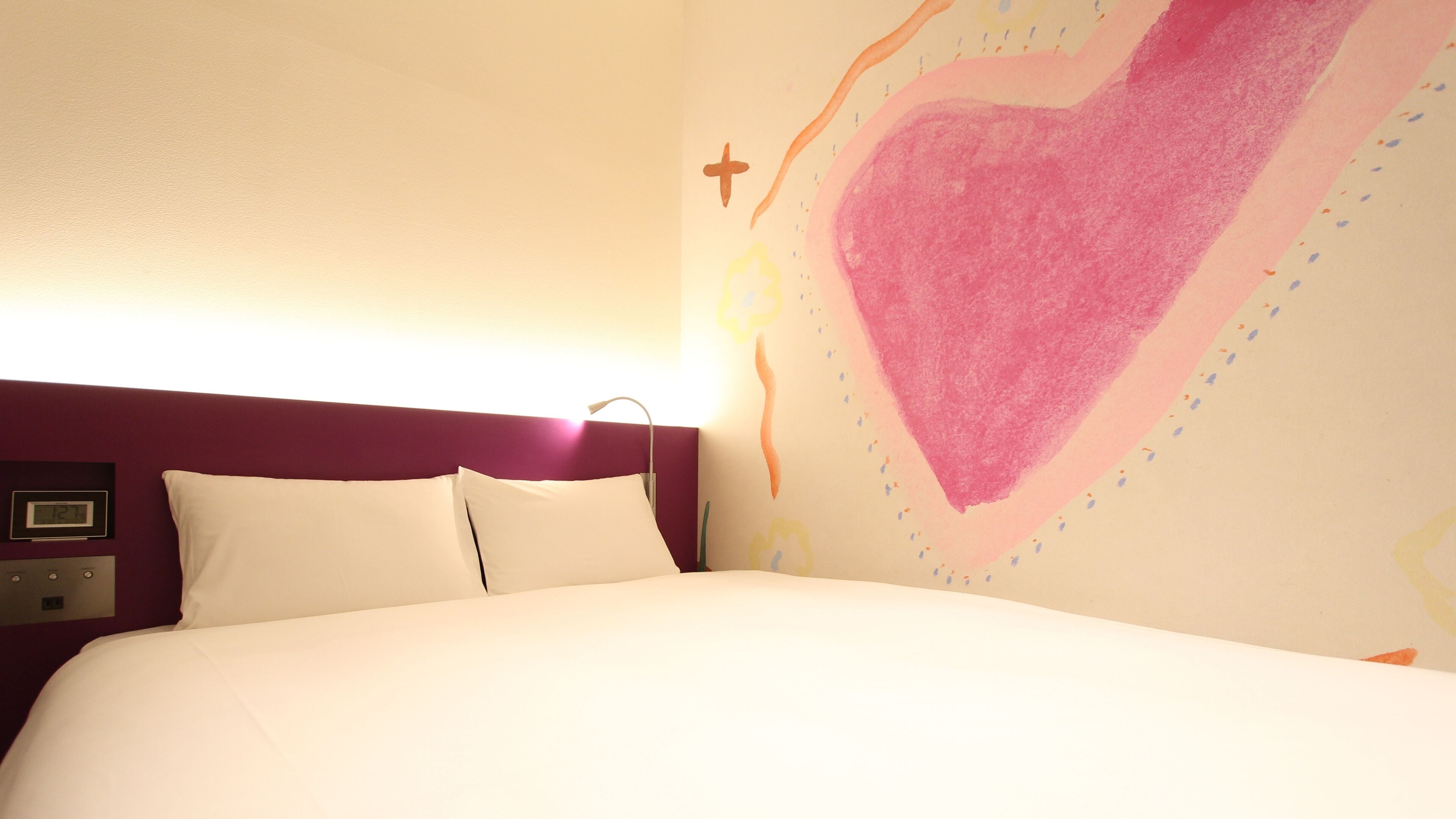 A bed with white bedding, a purple headboard, and a wall with a large heart design.