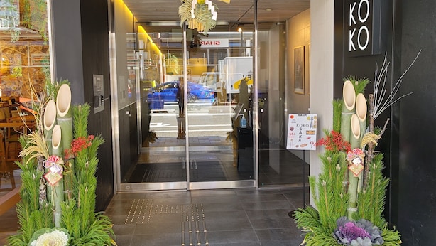 A entrada do Hotel Koko com design moderno, apresentando uma porta de vidro, uma placa de identificação e plantas decorativas.