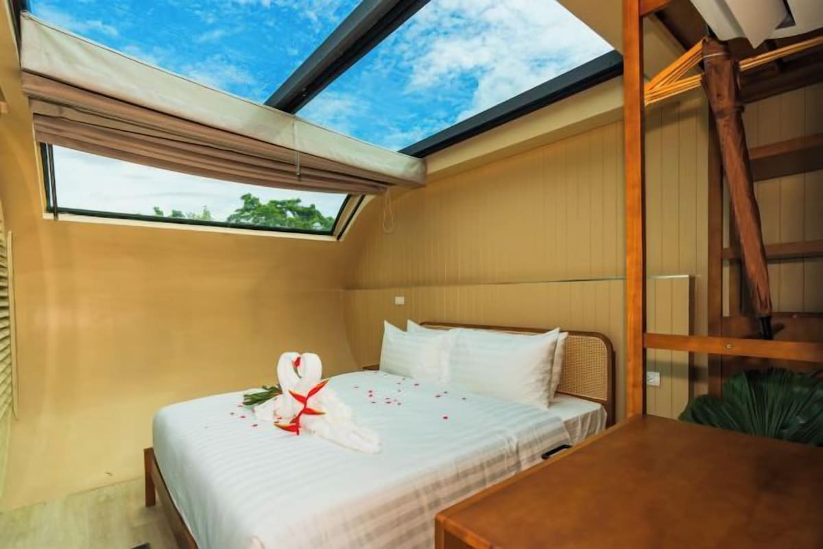 A single bed with a white bedspread and pillows, a wooden headboard, a skylight, and a wooden table.