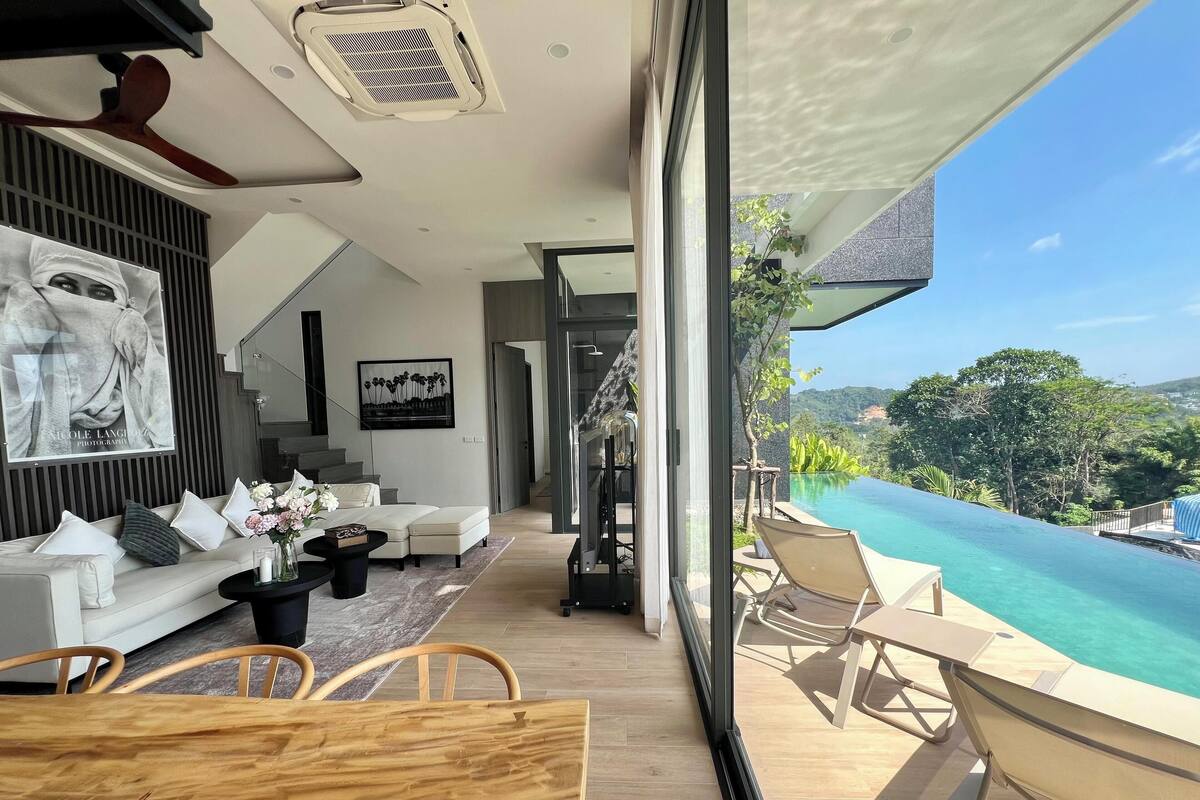 A modern living room with a sofa, coffee table, and chairs. There is a staircase leading to a balcony with a pool and a view of the mountains.