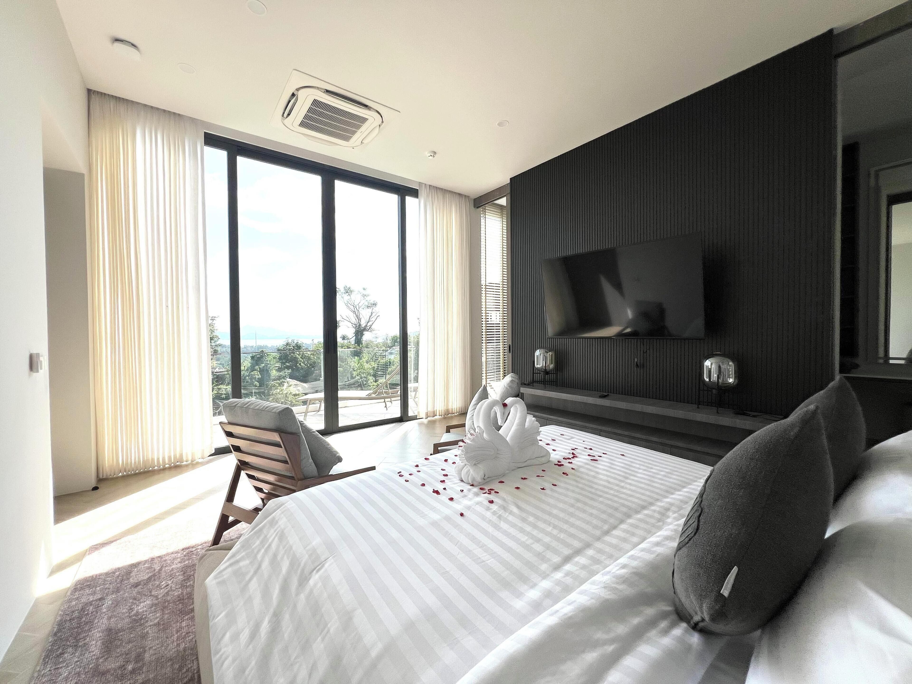 A modern hotel room with a large bed, a television mounted on the wall, and a view of the outdoors through the windows.