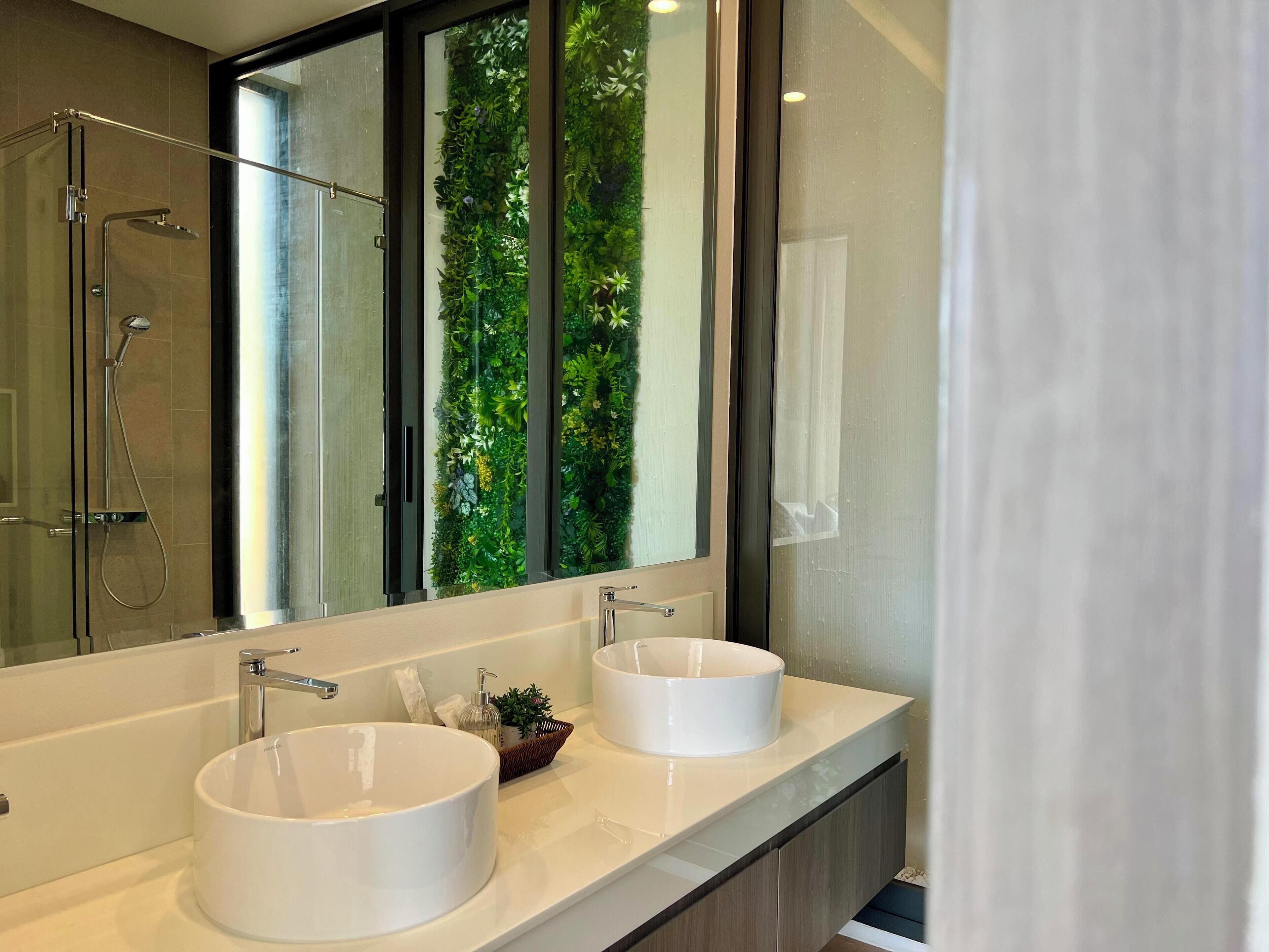 A modern bathroom with two white sinks, a glass shower enclosure, and a large mirror reflecting a green vertical garden.