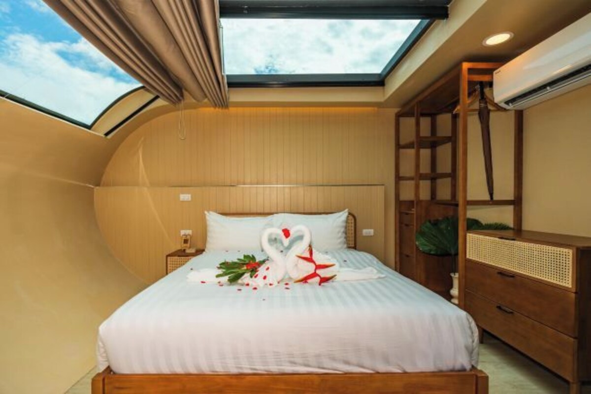 Un dormitorio con tragaluz, una cama con ropa de cama blanca, cabecera de madera, mesita de noche y cómoda.