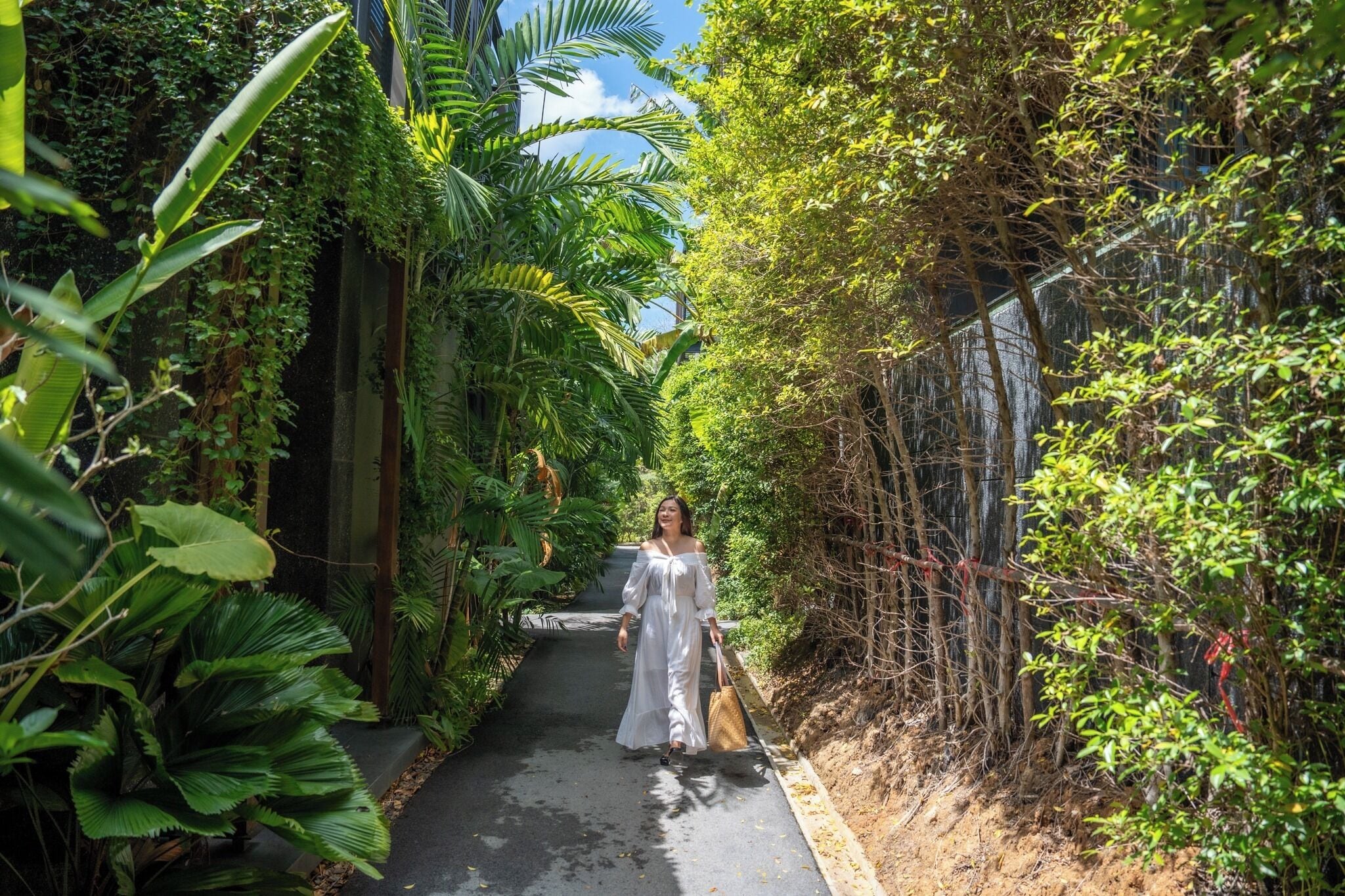 Un étroit sentier bordé d’une végétation luxuriante et d’une clôture en bambou, qui mène à un bâtiment.
