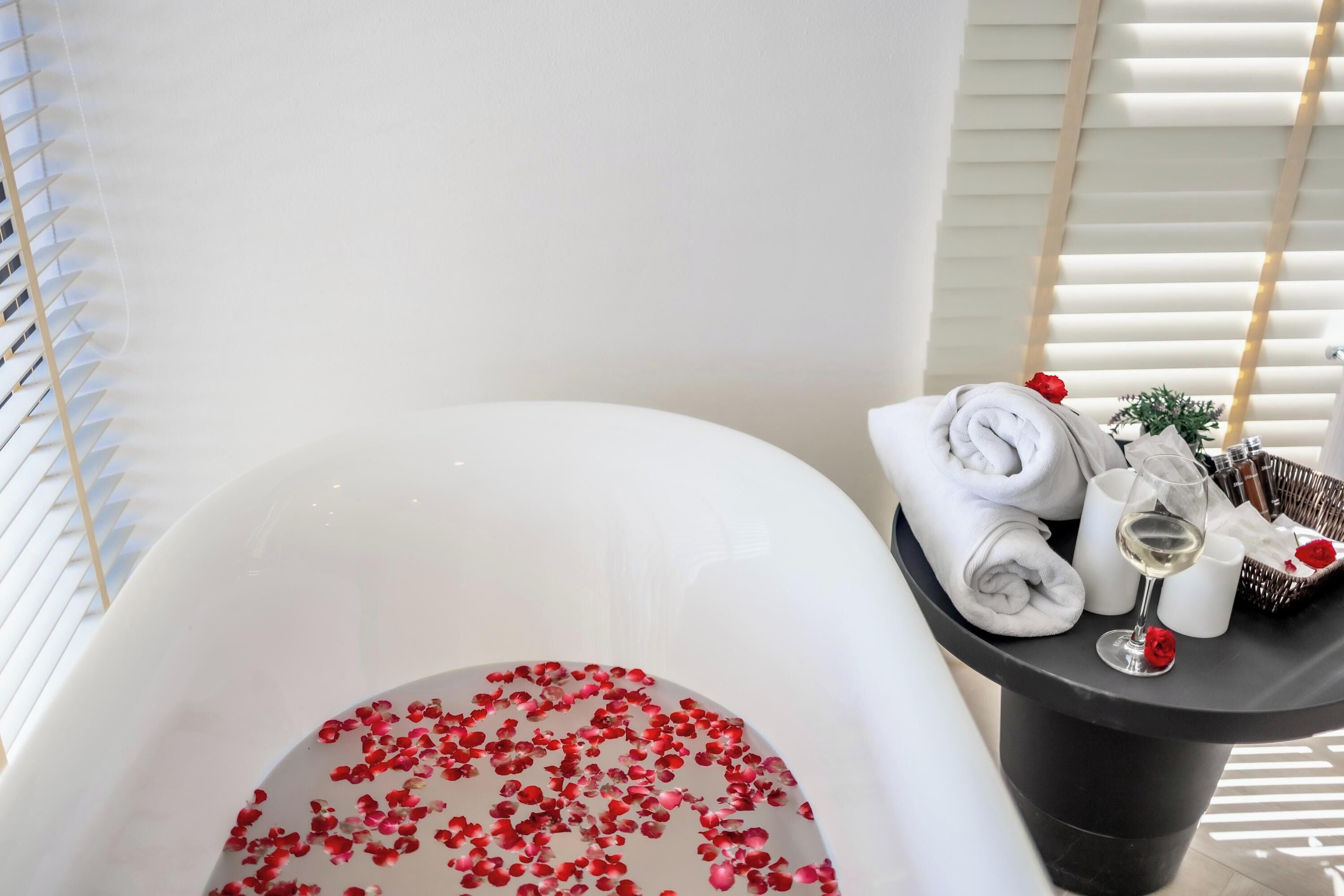 A bathtub filled with water and rose petals, a glass of white wine, and neatly folded towels on a side table.