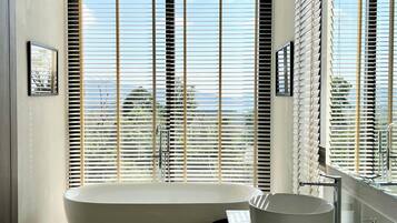 Een moderne badkamer met een vrijstaand bad, een wastafelmeubel en grote ramen met zonwering.