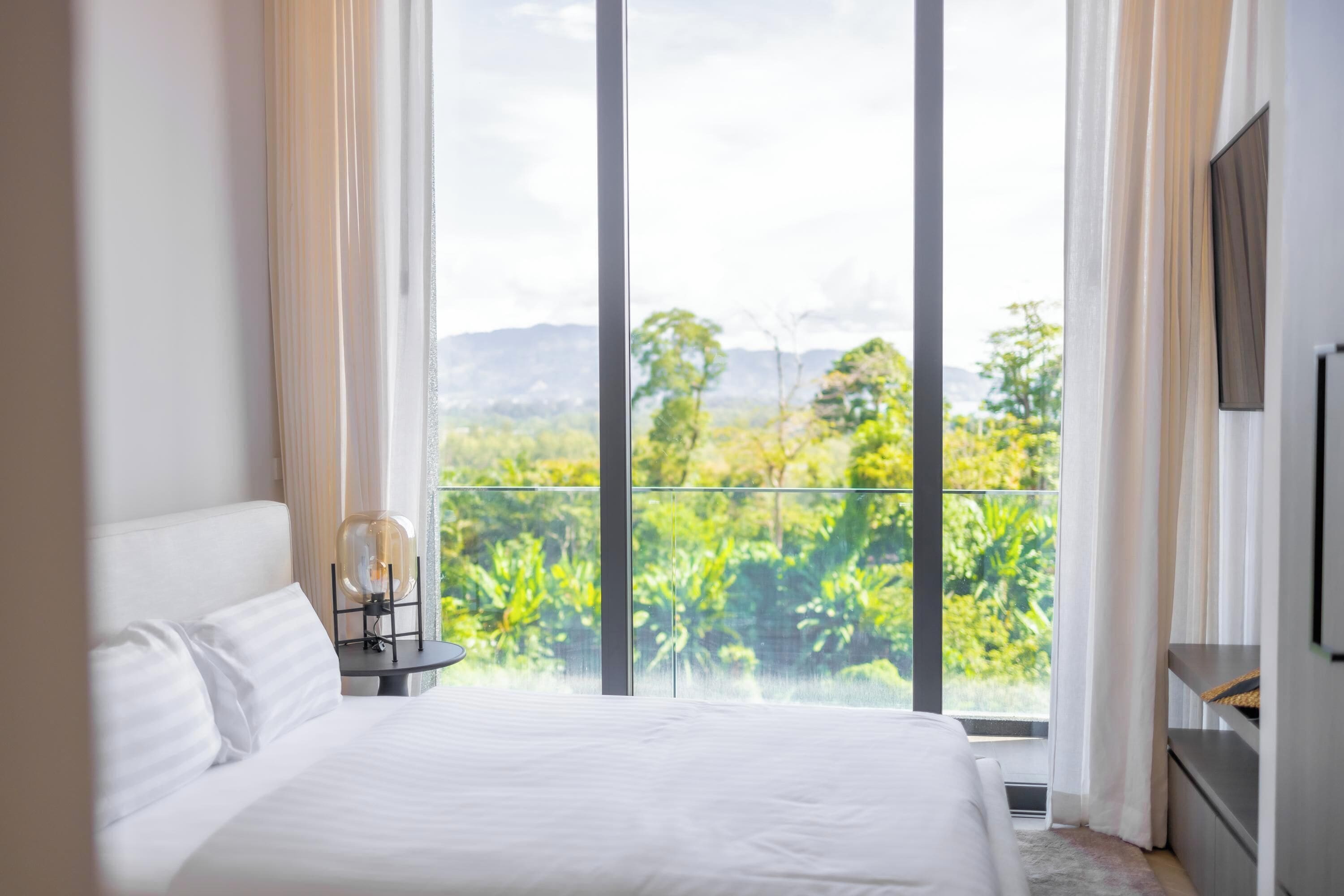 Ein Hotelzimmer mit einem großen Bett, einem Nachttisch, einem Fernseher und einem Balkon mit Blick auf Grünflächen und Berge.