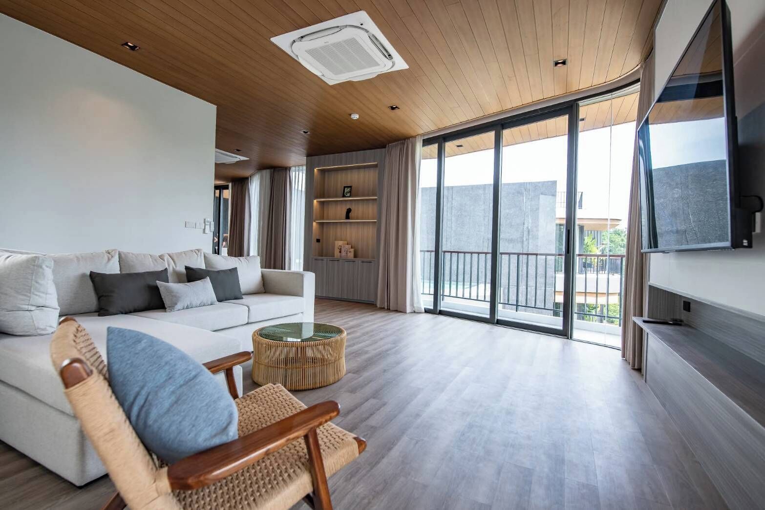 A spacious living room with a white sofa, a wooden chair, a glass coffee table, and a large window offering a view of the outdoors.