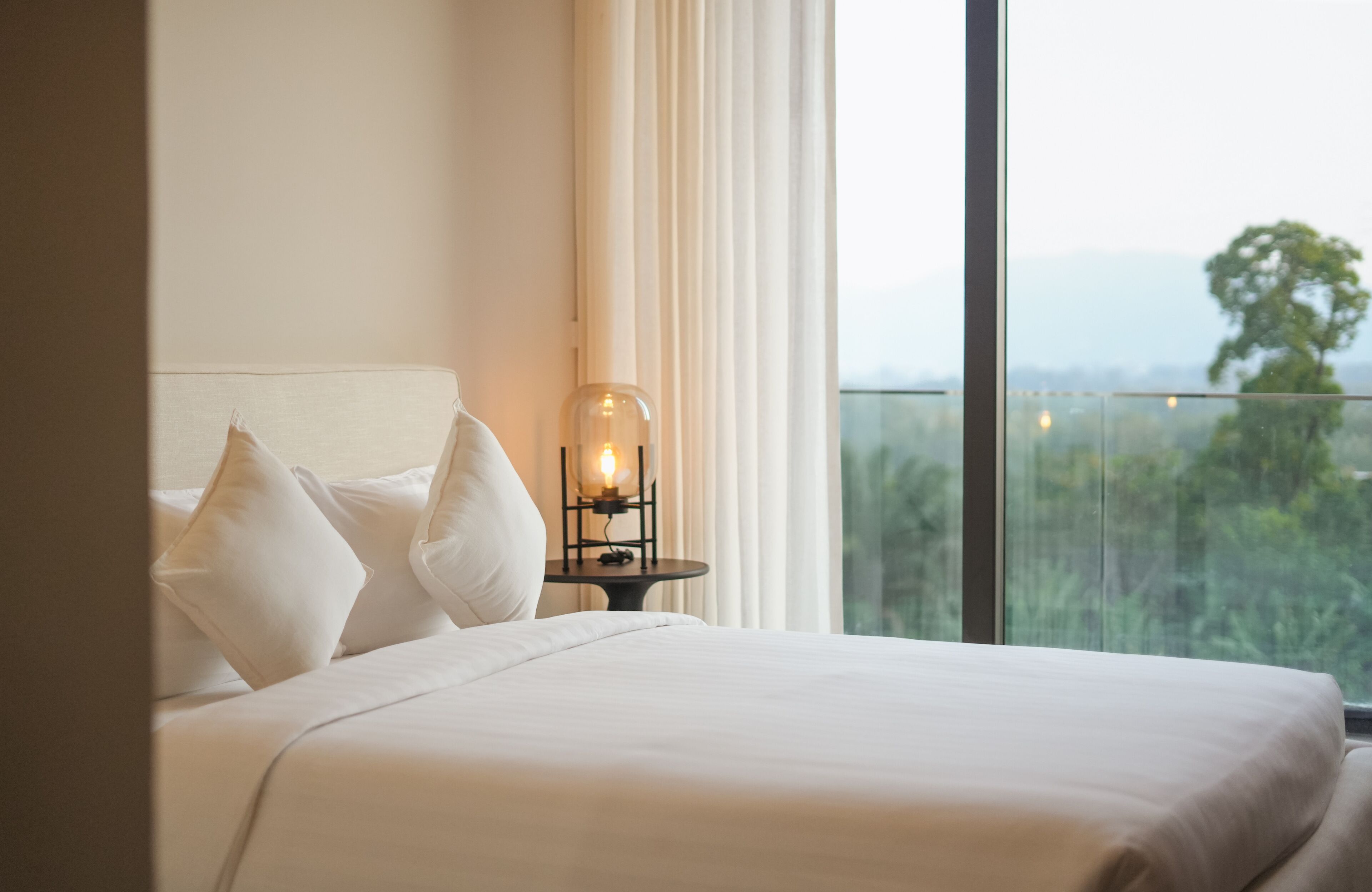 Una habitación de hotel con cama, ropa de cama blanca, una mesita de noche con lámpara y un ventanal con vistas a zonas verdes.