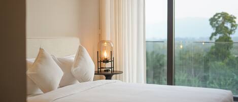 Una habitación de hotel con cama, ropa de cama blanca, una mesita de noche con lámpara y un ventanal con vistas a zonas verdes.