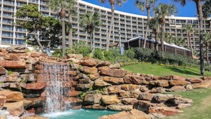 A hotel with a large building, a waterfall, and palm trees. - Gorgeous San Luis Condo! Amazing Pool & Amenities (Galveston)
