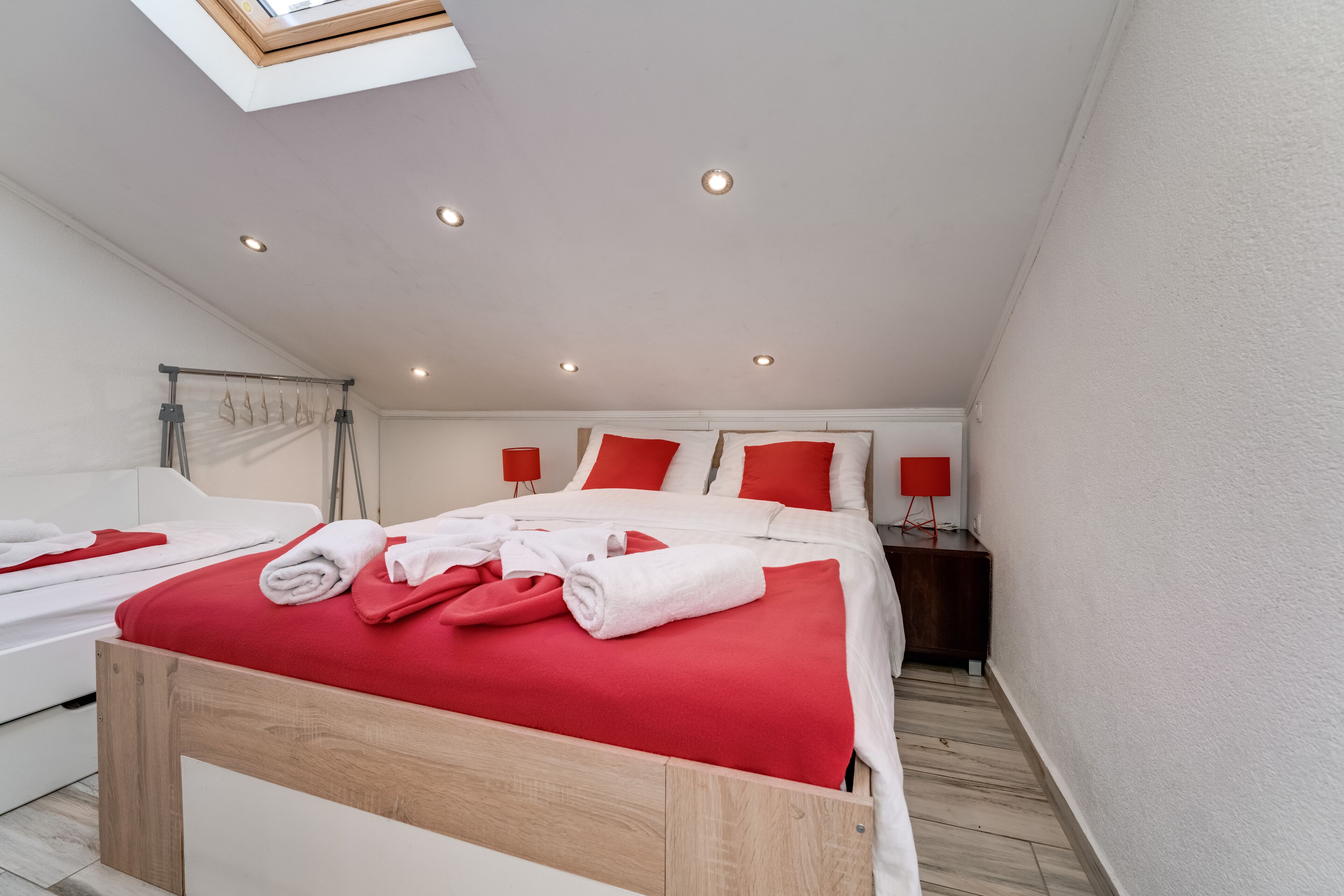 A modern bedroom with a skylight, a bed with red bedding, a bedside table with a red lamp, and a wooden headboard.