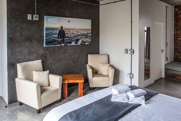 A modern hotel room with a large bed, two armchairs, a small wooden table, and a large poster of a person kite surfing.